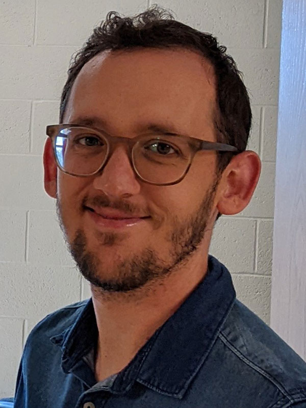 A headshot of Kody Steffy, who wears glasses and a dark shirt and poses against a white wall.