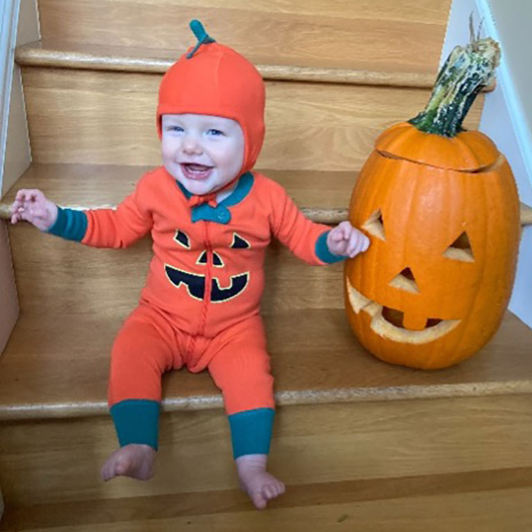 Dressed in a jack-o-lantern costume, a baby poses next to a pumpkin.