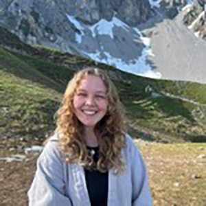A headshot of Myah Anne Clayborn, who poses in a jacket and black shirt with a mountain range in the background.