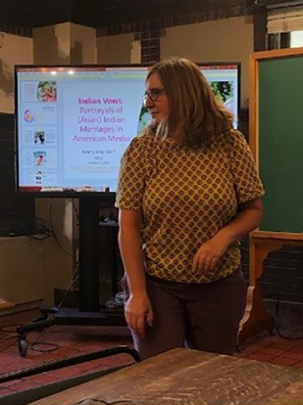 A photo of Keera Allendorf, who stands in front of a presentation on a television screen.