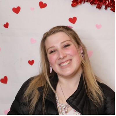 A headshot of Nora Barth, who poses against a white wall decorated with cartoon hearts.