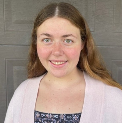 A headshot of Jenna Leigh Vander Wey, who wears a white sweater and poses against a gray background.