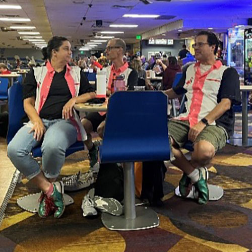 Three people in matching bowling shirts sit and talk at a bowling alley.