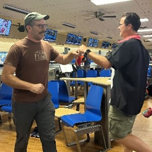 Two men smile and fist-bump near bowling lanes, with one wearing a casual t-shirt and the other in a bowling shirt.