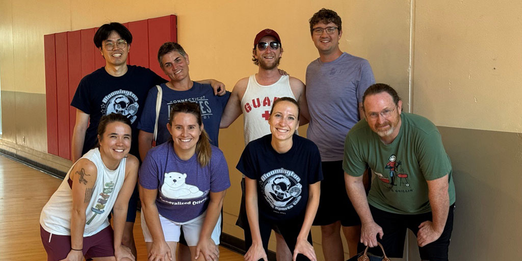 Eight people in gym clothes pose together in front of a tan wall and basketball court.