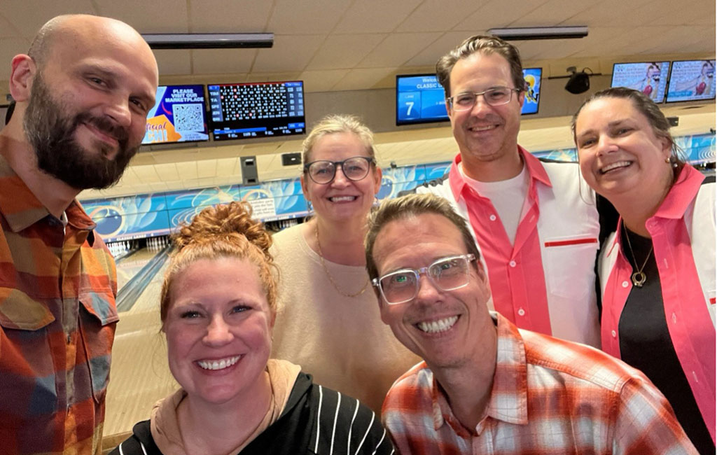 A group of six people, some in bowling shirts, smile for a selfie at the end of a bowling lane.