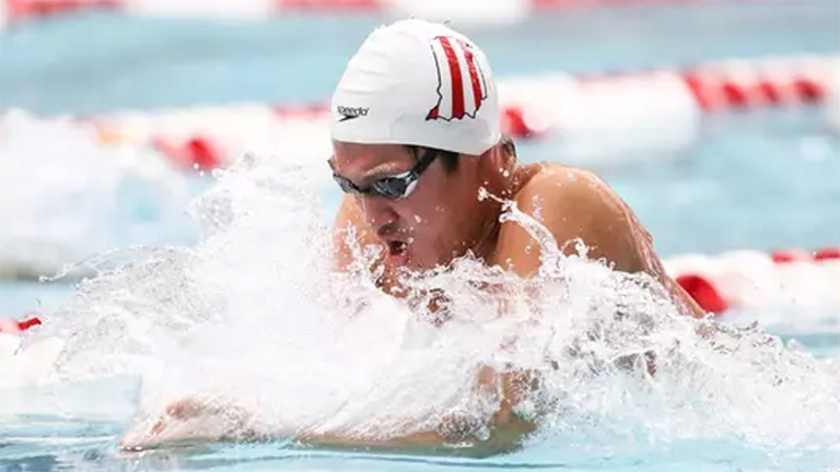 A competitive swimmer in a white IU swim cap powers through the water during a breaststroke race.