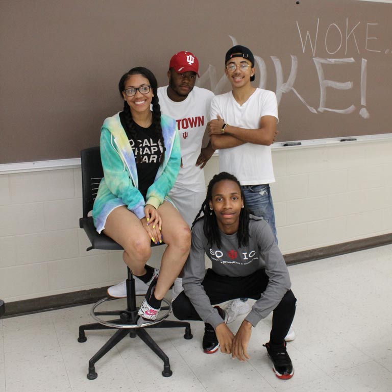 Four students pose in front of a chalkboard that says, "WOKE"